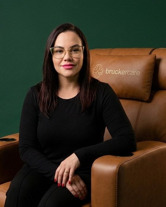 Pâmela Zweibrucker, fundadora da BruckerCare, sentada numa poltrona de couro caramelo, de blusa preta e óculos, sorrindo para a câmera.